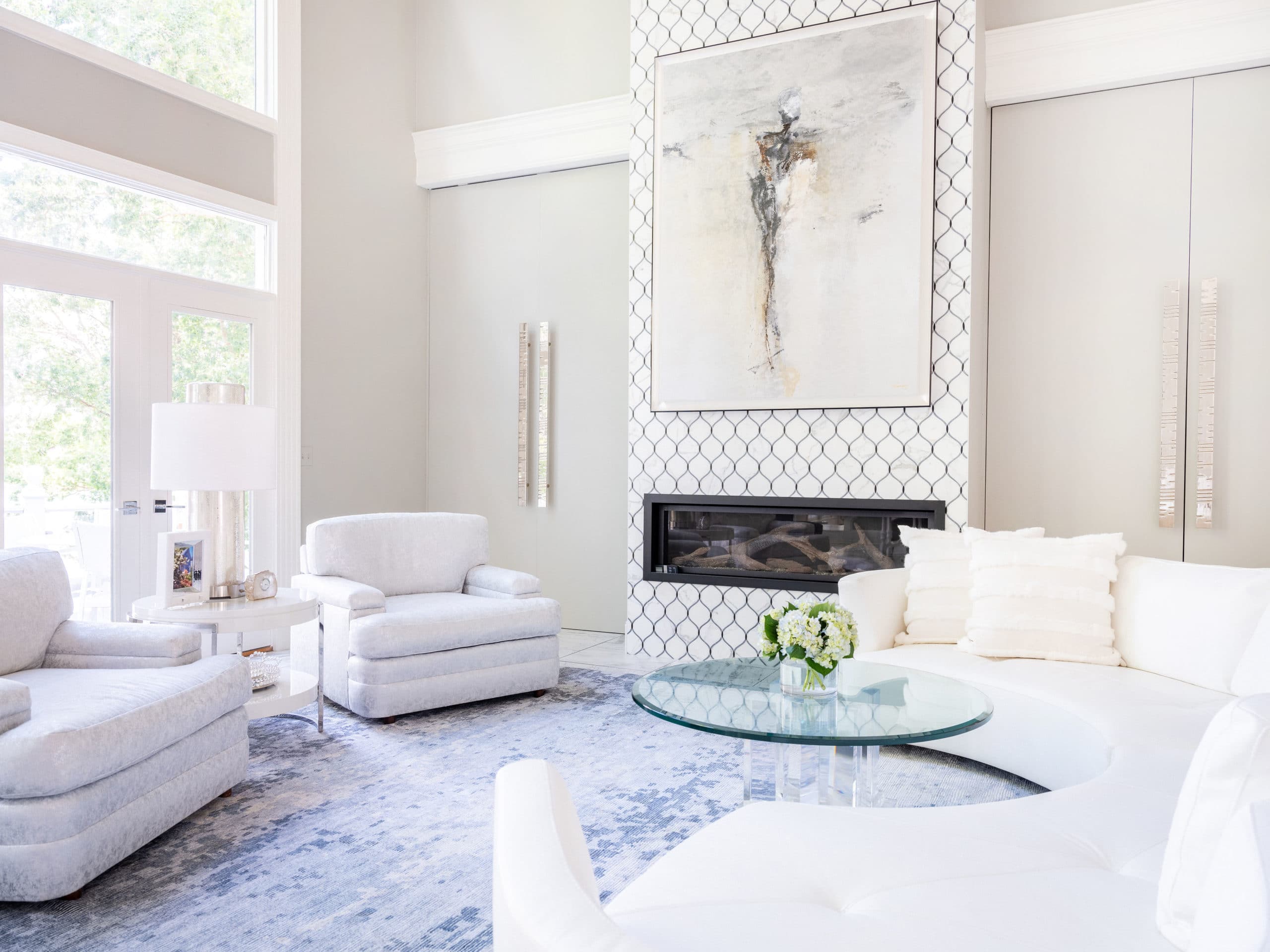 All white living room filled with couches, chairs, fireplace, glass coffee table, fireplace, and large picture.